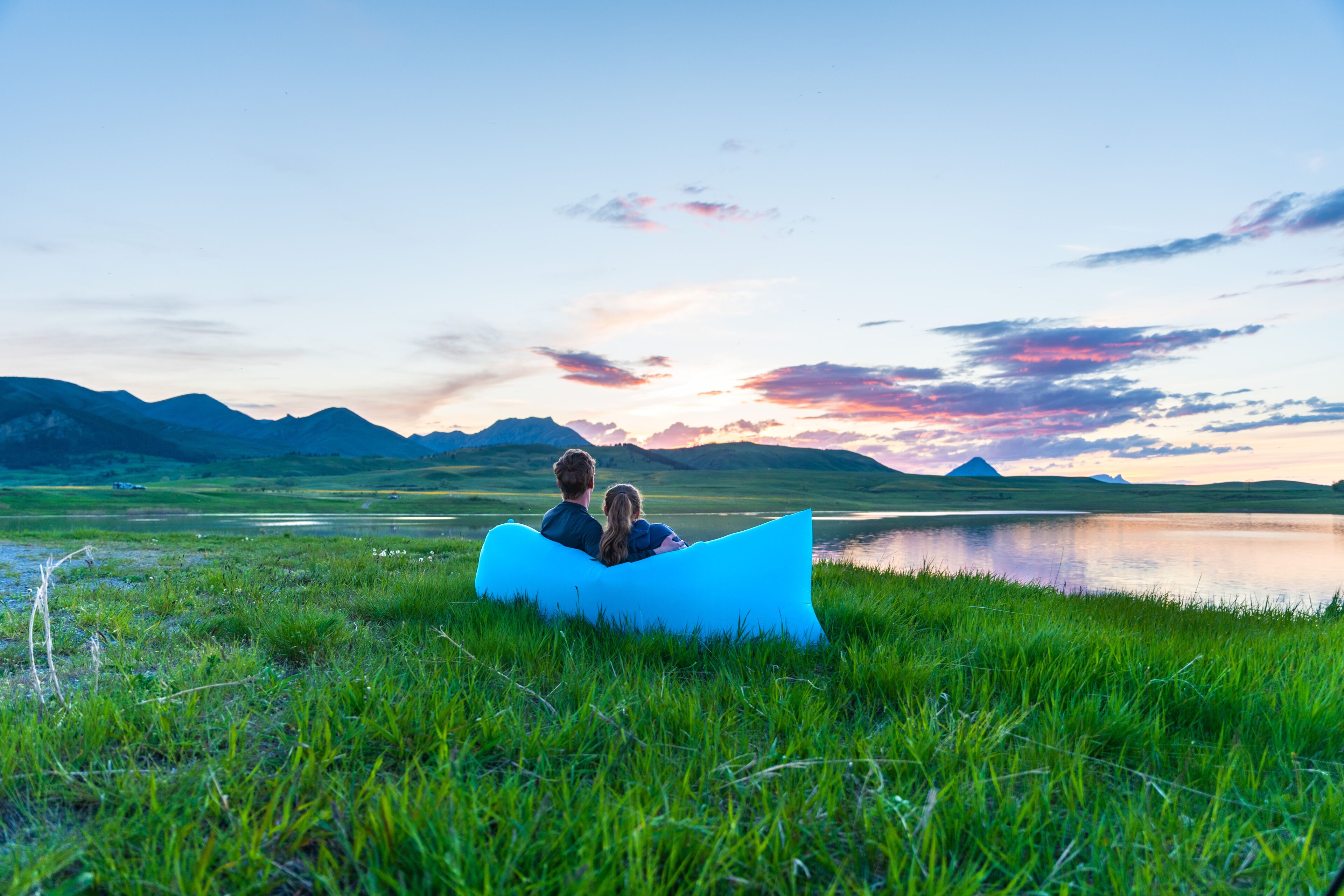 Una pareja en un canoa azul sobre un campo cubierto de hierba, con un lago y montañas al fondo.