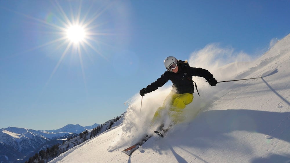 Un skieur, vêtu d’un pantalon jaune et d’une veste noire, descend une pente enneigée, avec des montagnes en arrière-plan et un soleil brillant.