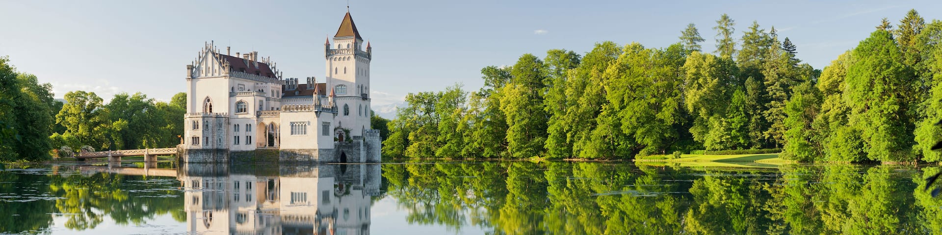 A castle with a tower, surrounded by a forest and reflecting in a calm lake.