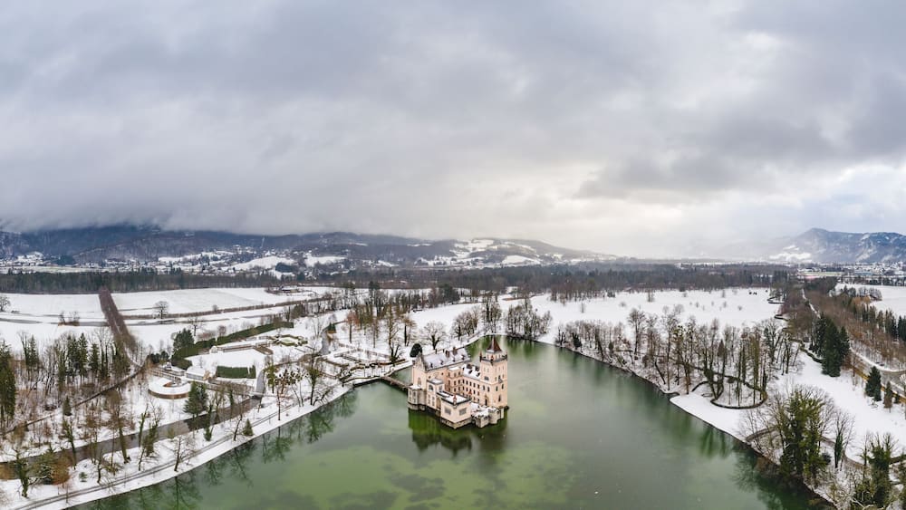 Un paysage enneigé avec un château au bord d’une rivière, des montagnes en arrière-plan et un ciel nuageux.