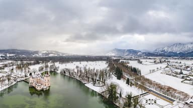 Paisagem coberta de neve com um castelo à beira do rio, montanhas ao fundo e céu nublado.