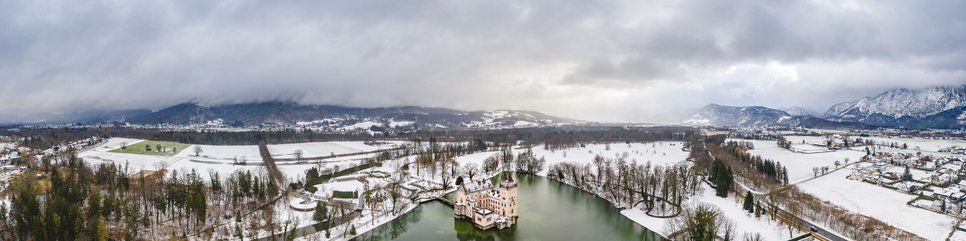 Paisaje nevado con un castillo junto a un río, montañas al fondo y cielo nublado.