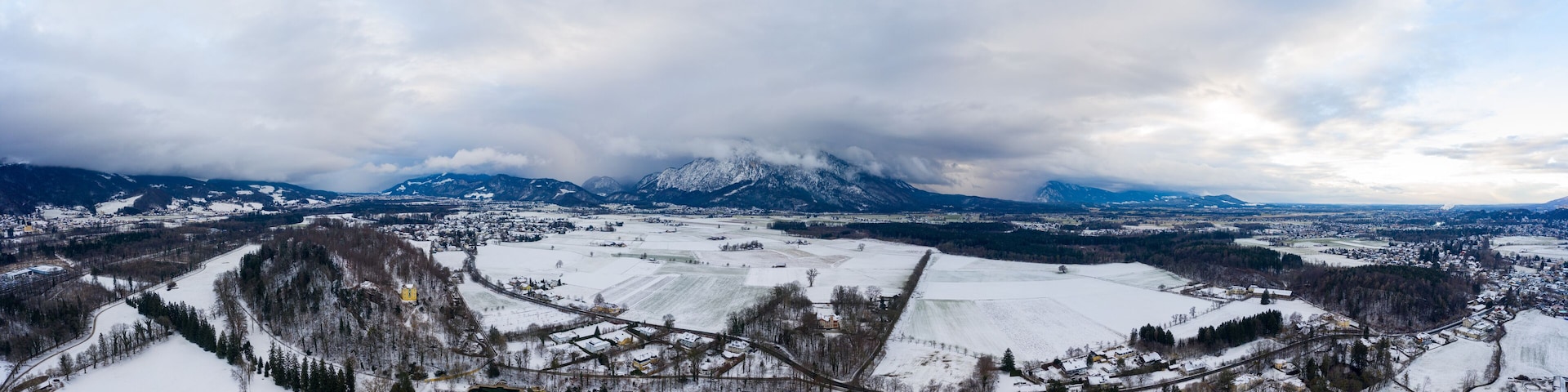 A panoramic view of a snow-covered landscape with mountains in the background, a town in the foreground, and a railway track.