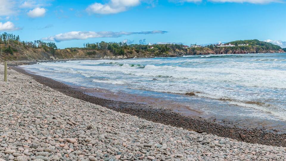 A pebbled beach with waves, a forested hill, and houses in the distance.