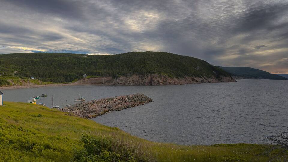 A coastal landscape with a house, a rocky breakwater, and a forested hill under a cloudy sky.