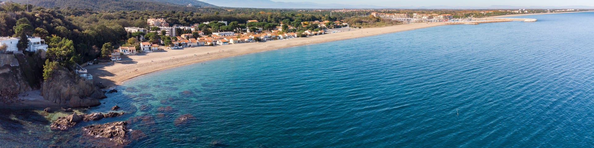 A coastal town with a beach, clear blue sea, and mountains in the background.