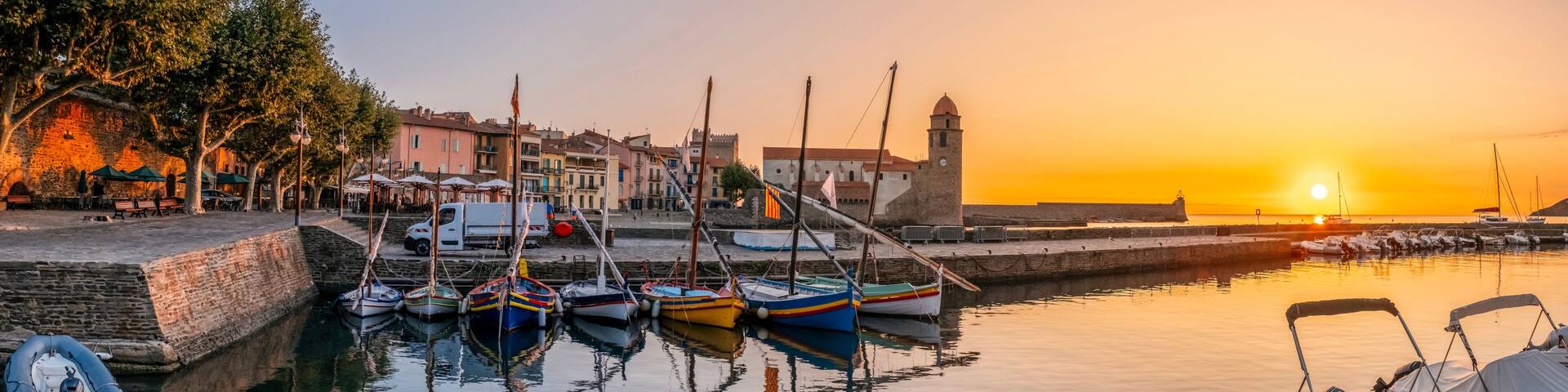 A harbor at sunset with boats docked, a stone wall, and buildings in the background.