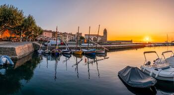 A harbor at sunset with boats docked, a stone wall, and buildings in the background.