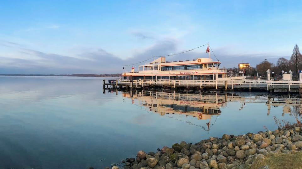 A calm lake with a large boat docked at the pier, surrounded by rocks and sparse vegetation.