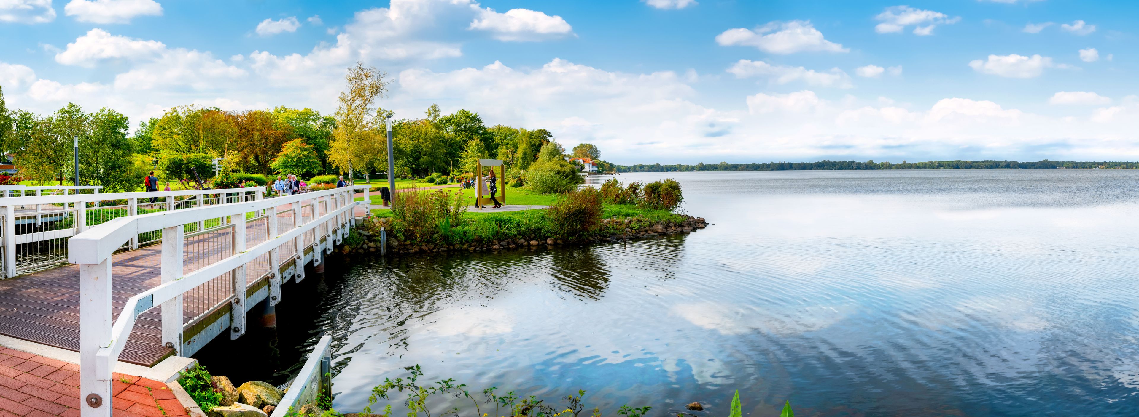 A white bridge over calm water, surrounded by lush greenery and a clear sky.