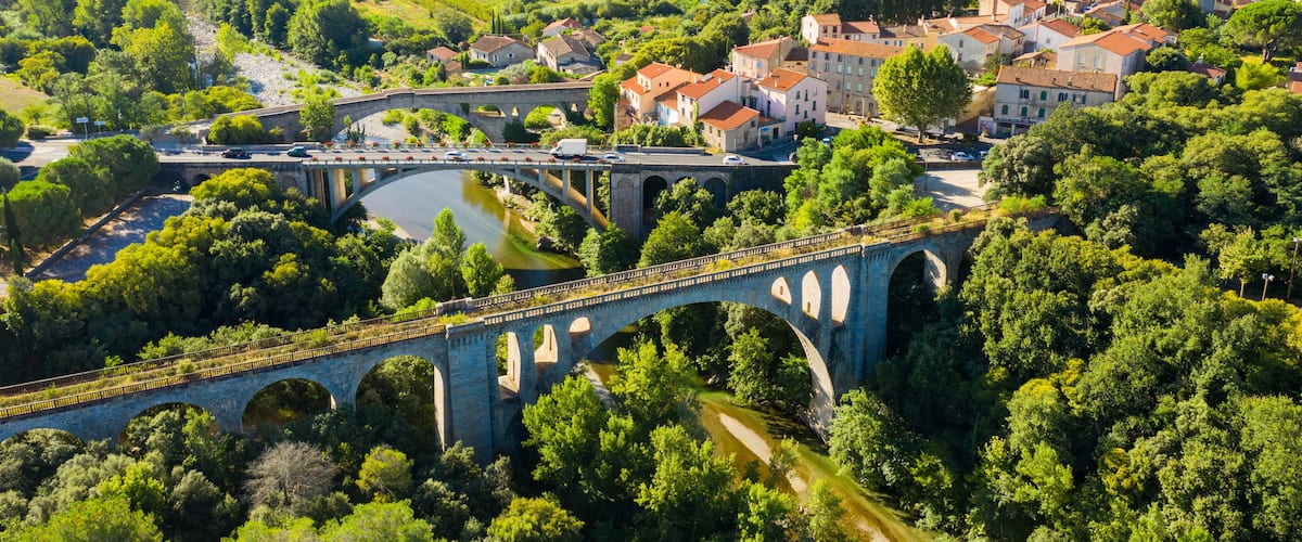 A stone bridge over a river, surrounded by lush greenery and a town with red-roofed buildings.