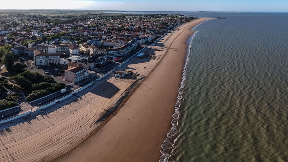 Une vue aérienne d’une ville côtière, avec une longue plage de sable, une zone résidentielle densément peuplée et une vue dégagée sur l’océan.