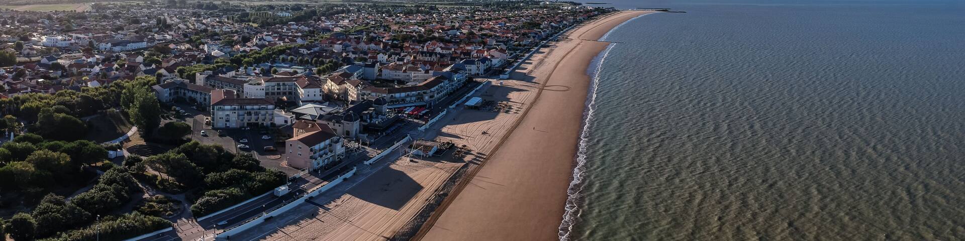 An aerial view of a coastal town with a long sandy beach, dense residential area, and a clear view of the ocean.