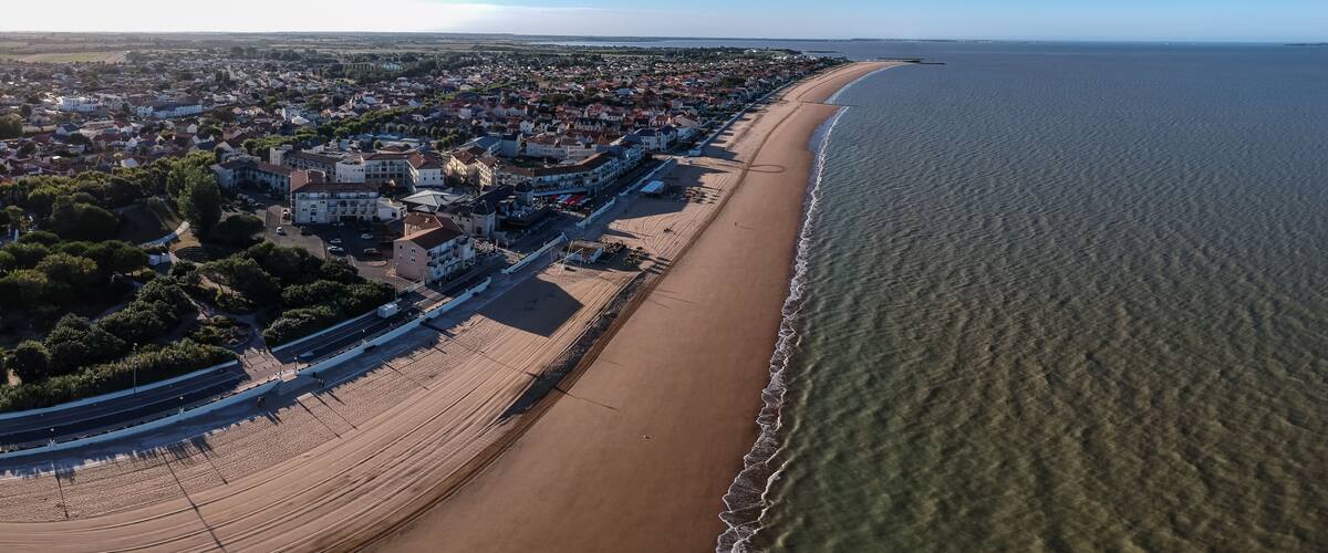 An aerial view of a coastal town with a long sandy beach, dense residential area, and a clear view of the ocean.