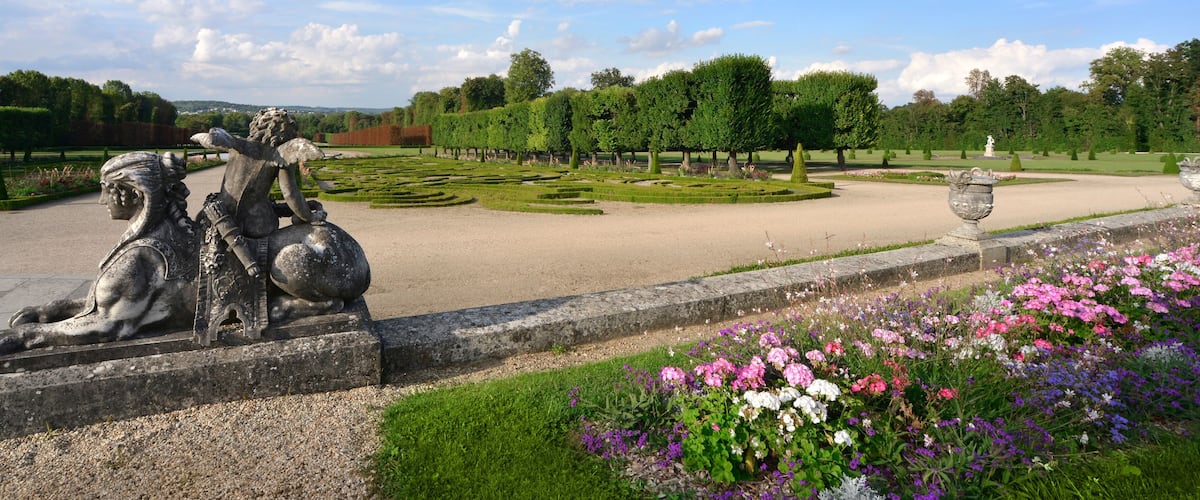 A statue of a seated figure with a child, surrounded by a formal garden with neatly trimmed hedges and flower beds.