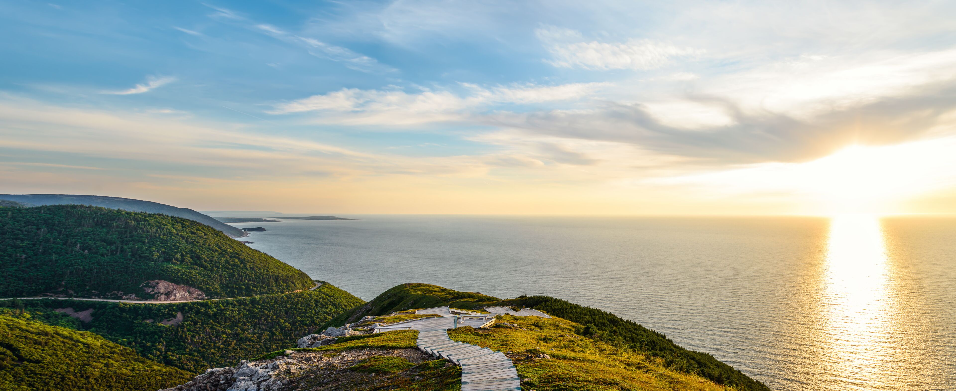 A coastal landscape with a winding path, lush green hills, and a calm sea under a clear sky.