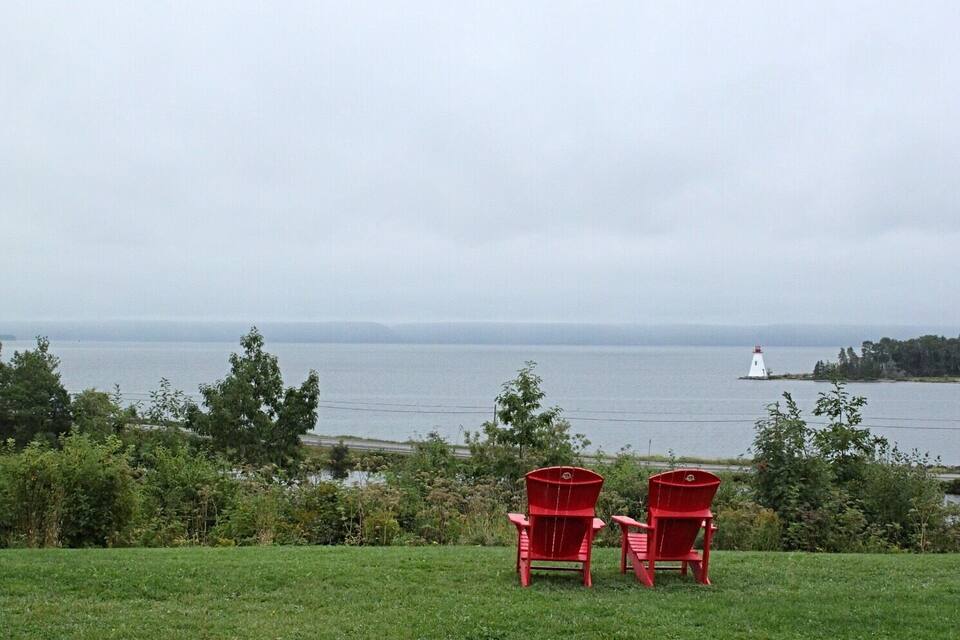 Two red Adirondack chairs facing a body of water with a lighthouse in the distance.