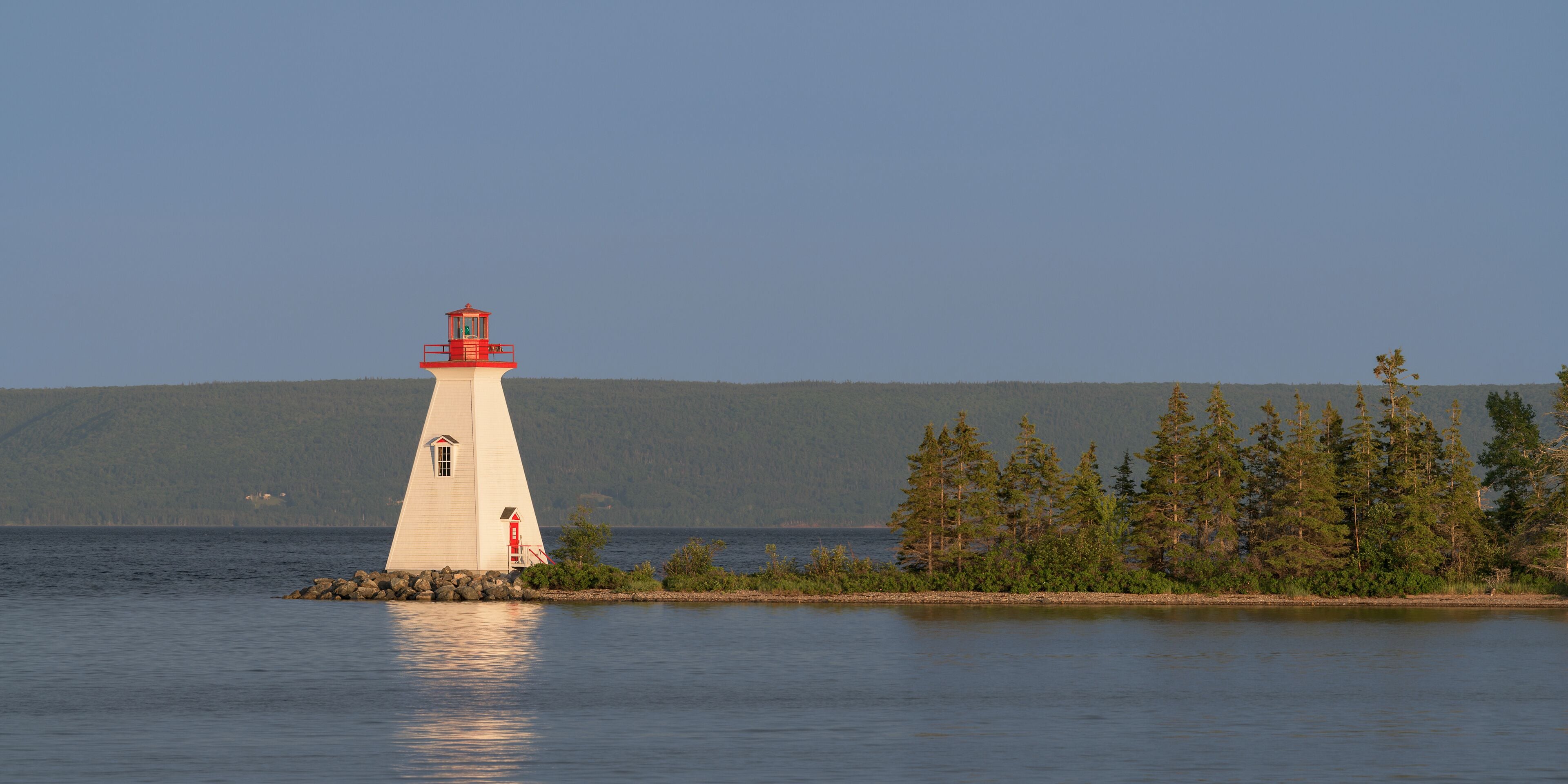 Un faro blanco con la parte superior roja en una pequeña isla rodeada de aguas tranquilas y un bosque denso.