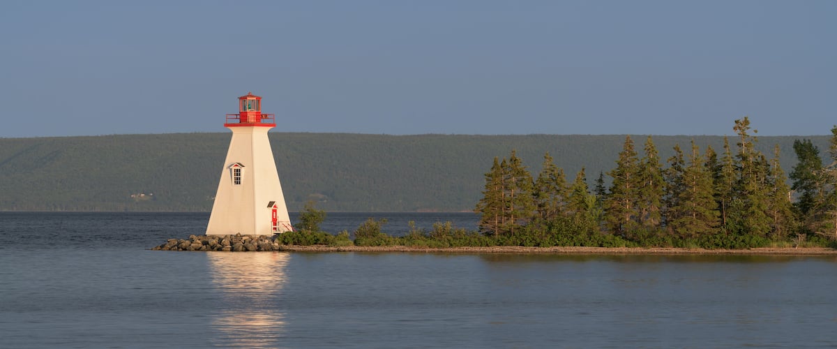 Un phare blanc, coiffé d’un dôme rouge, se dresse sur une petite île entourée d’eaux calmes et d’une forêt dense.