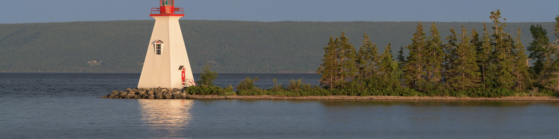 A white lighthouse with a red top on a small island surrounded by calm water and dense forest.