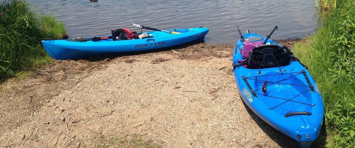 Two blue kayaks on a gravel shore by a calm lake with a distant hilly landscape.
