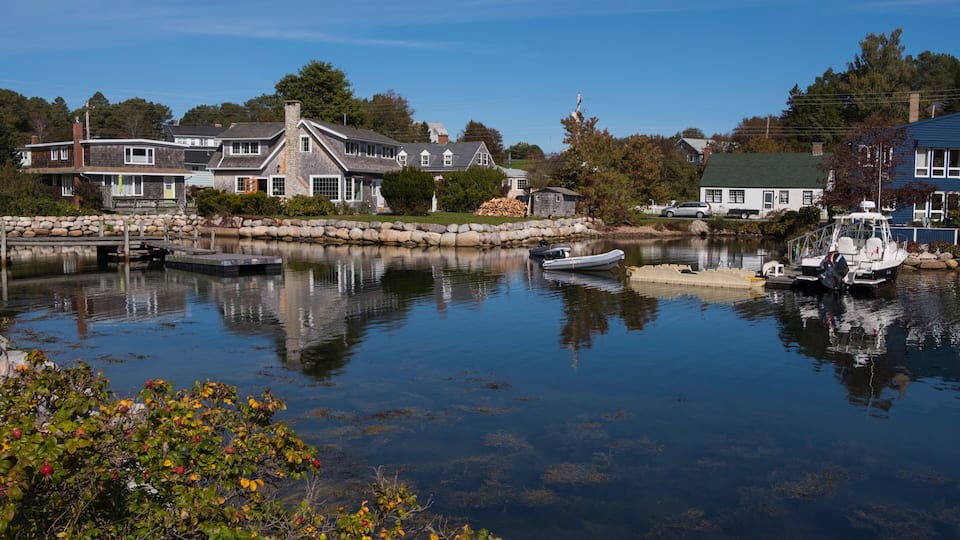 A calm lake with boats, a stone dock, and houses with a stone wall and trees in the background.