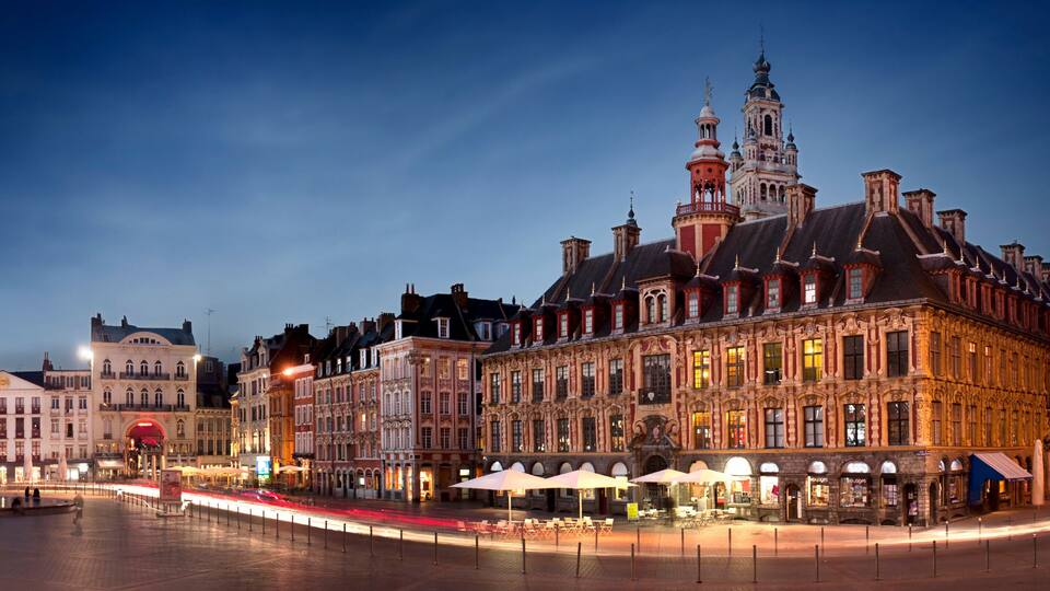 A historic European city square at dusk with illuminated buildings and a prominent clock tower.
