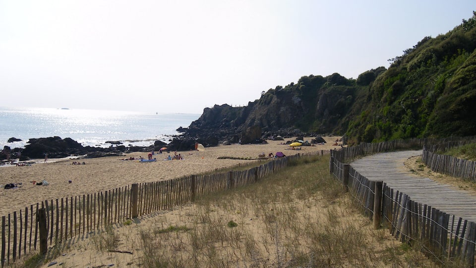 A sandy beach with a wooden boardwalk, rocky cliffs, and the ocean in the background.