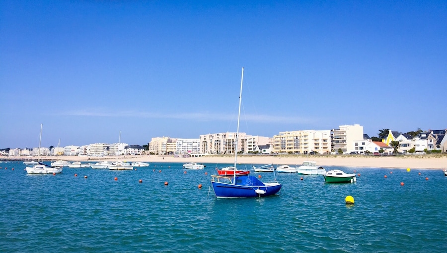 A coastal marina with various boats, a sandy beach, and buildings in the background.