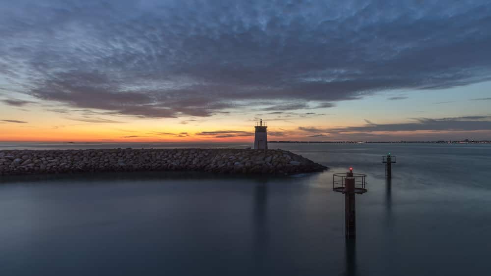 Eine Küstenlandschaft mit einem Leuchtturm, einem Stegabriss und ruhigem Wasser bei Sonnenuntergang.
