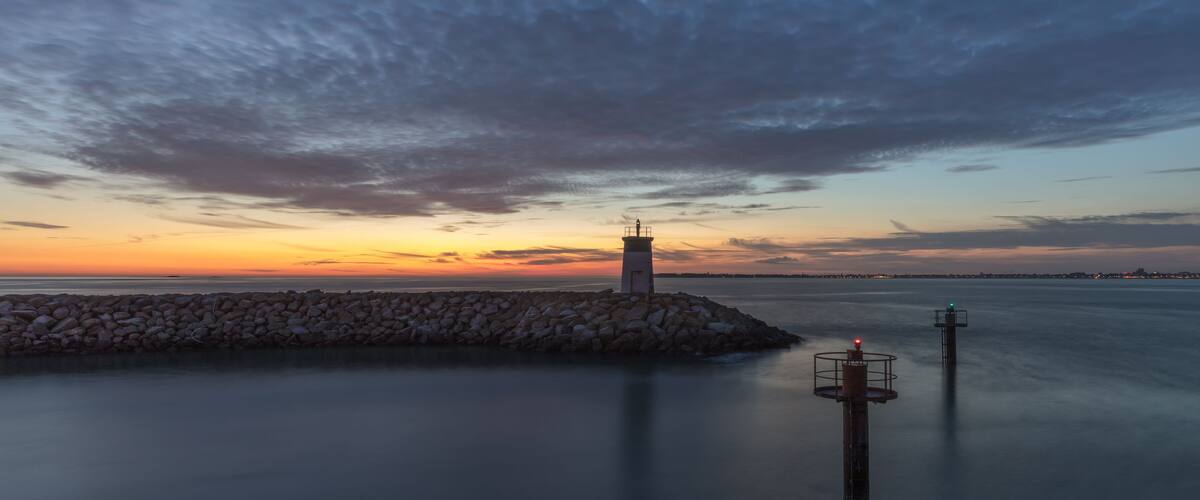 Een kustlandschap met een vuurtoren, een stenen golfbreker en rustig water tijdens zonsondergang.