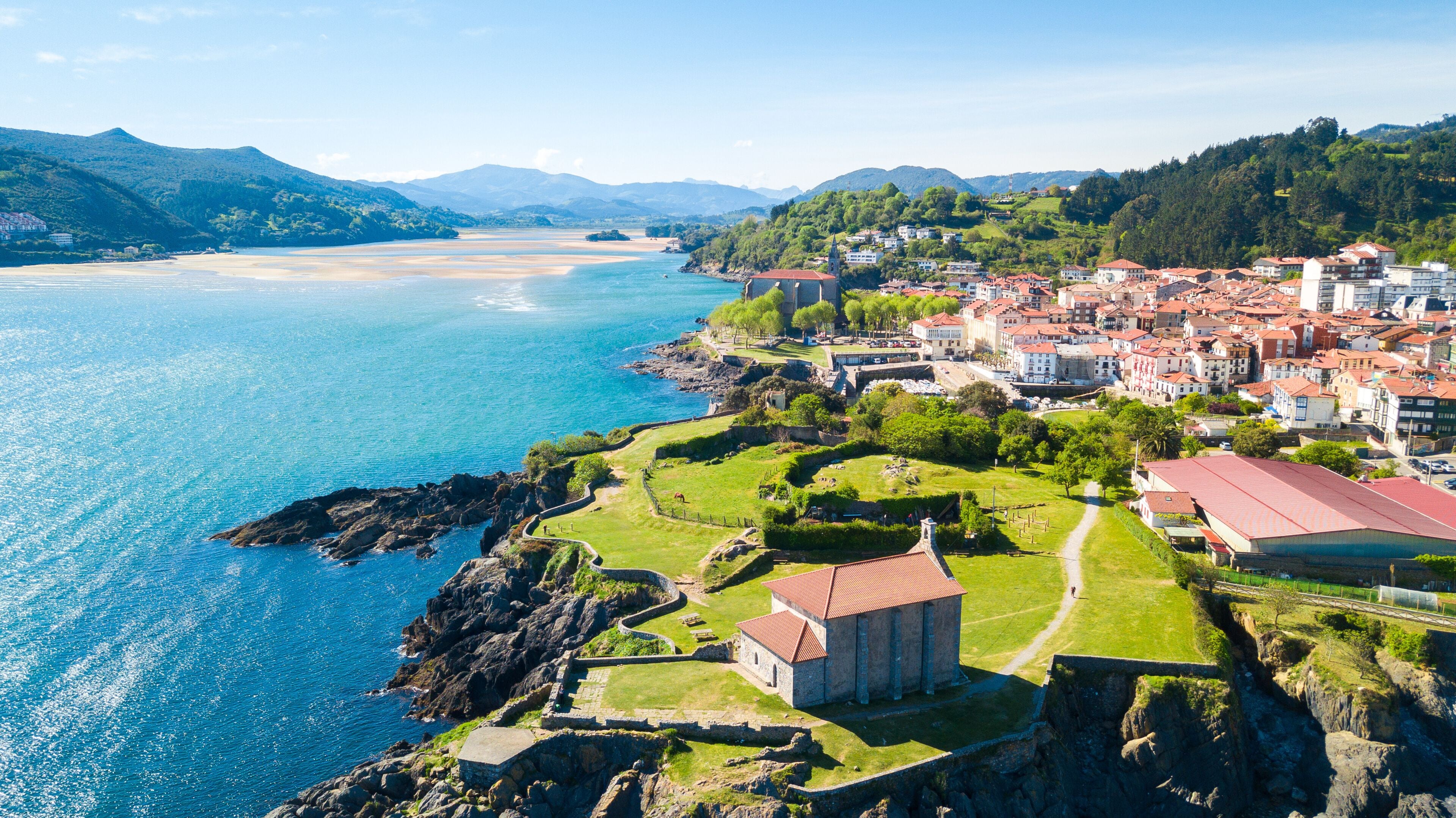 Uma cidade costeira com uma igreja histórica, uma praia e terreno montanhoso ao fundo.
