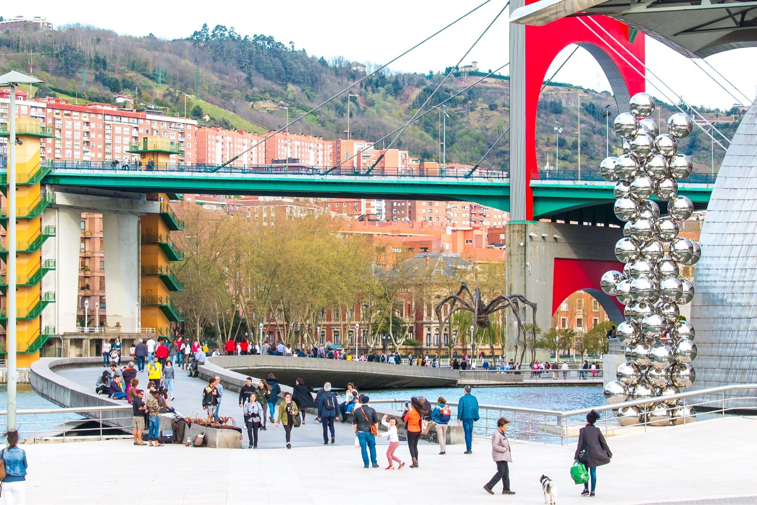 A bridge with a red arch, a sculpture of metallic spheres, and a waterfront promenade with people.