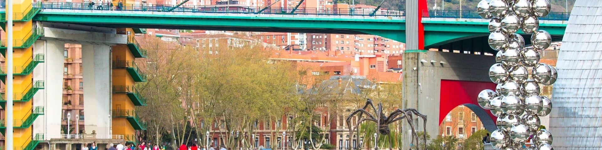 A bridge with a red arch, a sculpture of metallic spheres, and a waterfront promenade with people.