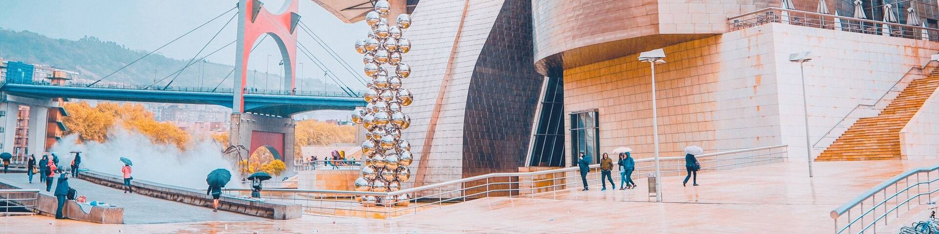 A modern architectural building with a unique curved design, a large metallic sculpture, and a bridge in the background.