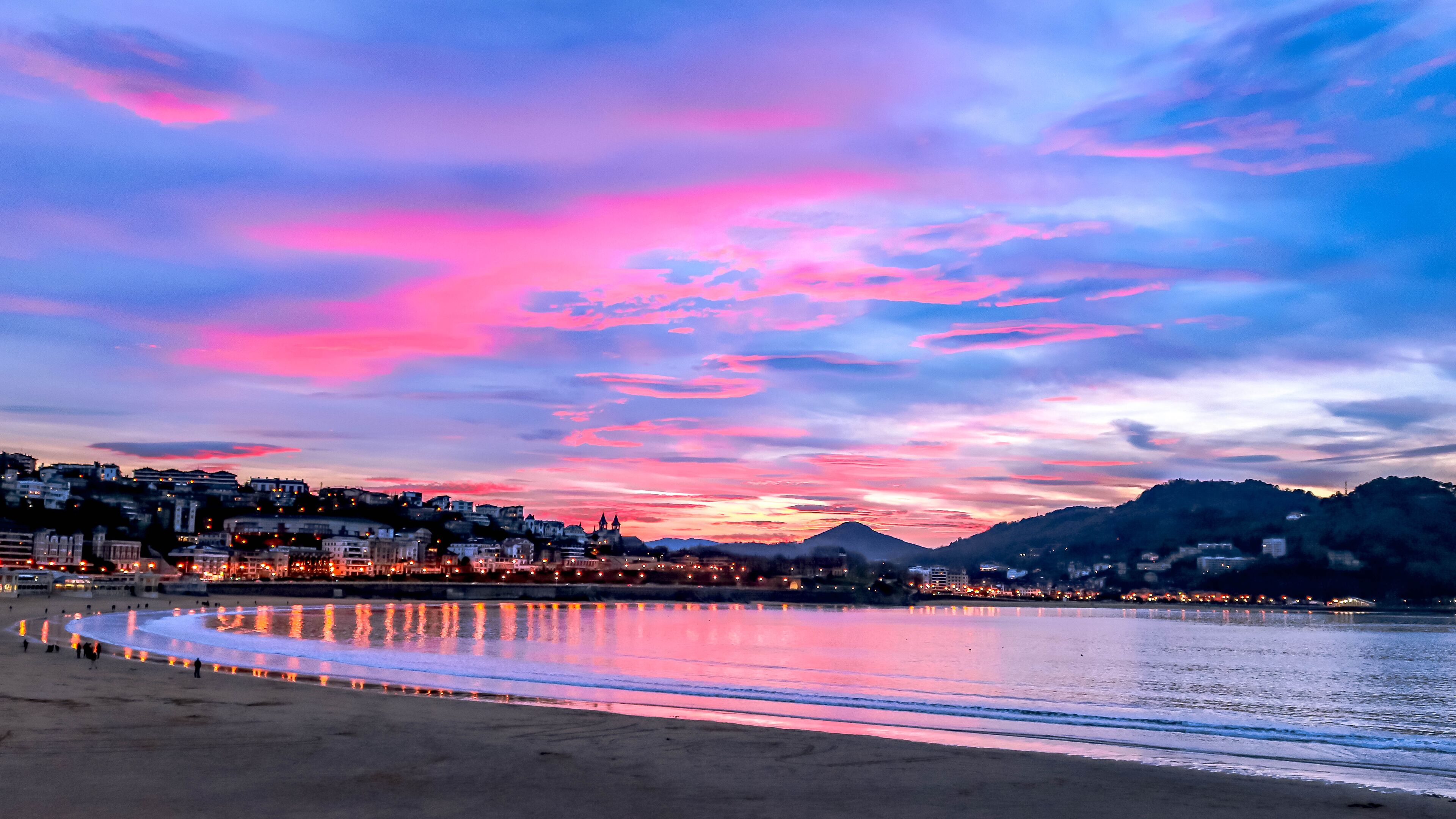 Uma praia com céu ao pôr do sol, águas calmas refletindo luzes e uma paisagem urbana ao fundo.