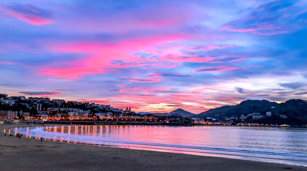 Uma praia com céu ao pôr do sol, águas calmas refletindo luzes e uma paisagem urbana ao fundo.