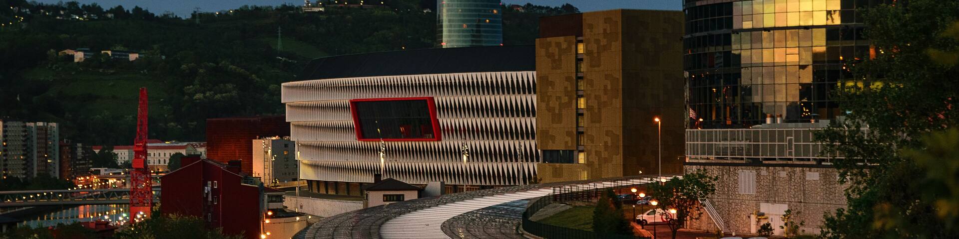 A modern cityscape with a curved road, a red building, and a glass skyscraper reflecting the sky.
