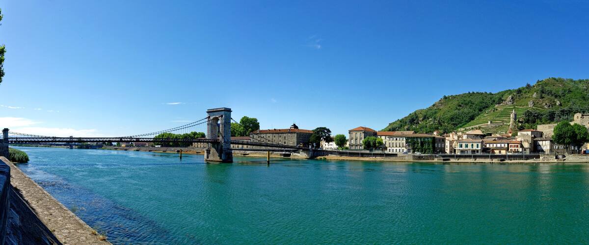A suspension bridge over a river, surrounded by buildings and a hillside.