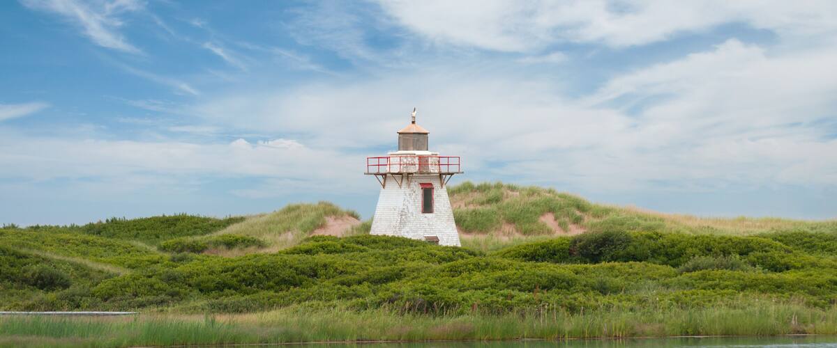 Close-up view of the lighthouse in St. Peter Harbour, Prince Edward Island, Canada, standing on sand dunes covered with green grass, reflecting on the water.