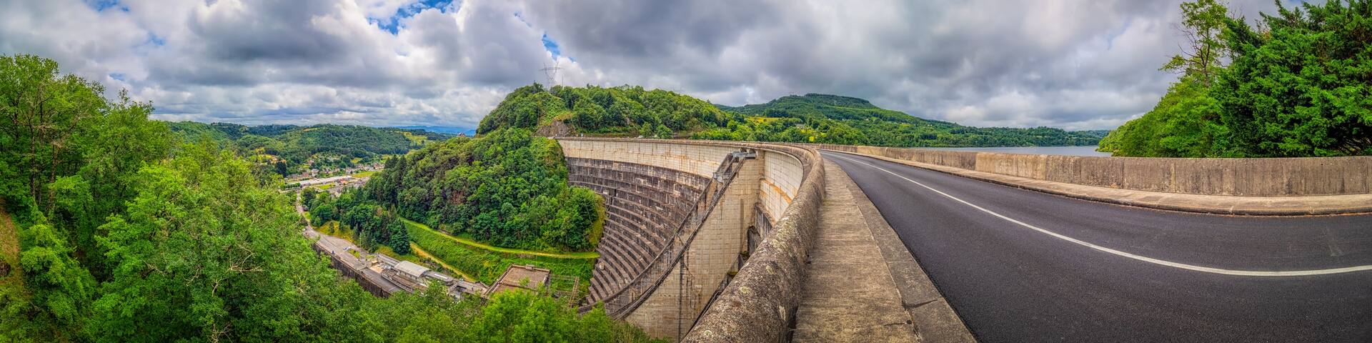 A dam with a road curving alongside it, surrounded by lush greenery and hills.