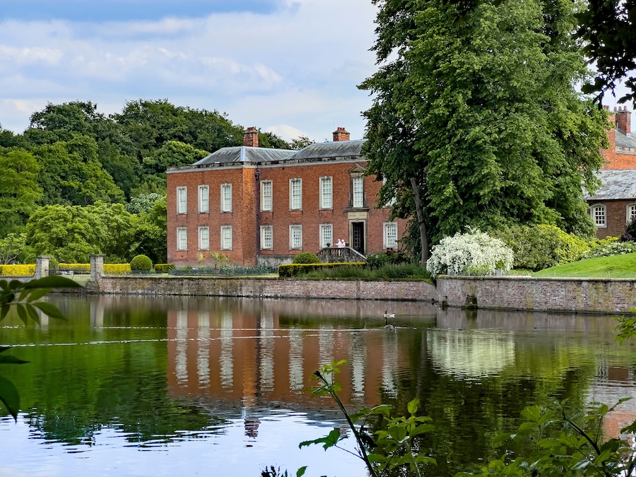 A large brick building with a pond in front, surrounded by trees and greenery.