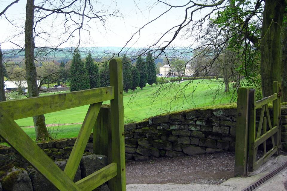 A stone wall and wooden gate leading to a green field with trees and a distant building.