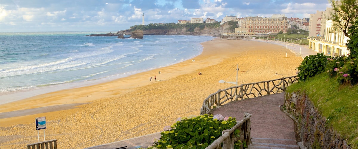 A coastal beach with a stone staircase leading down to the shore, overlooking the ocean and a row of buildings.