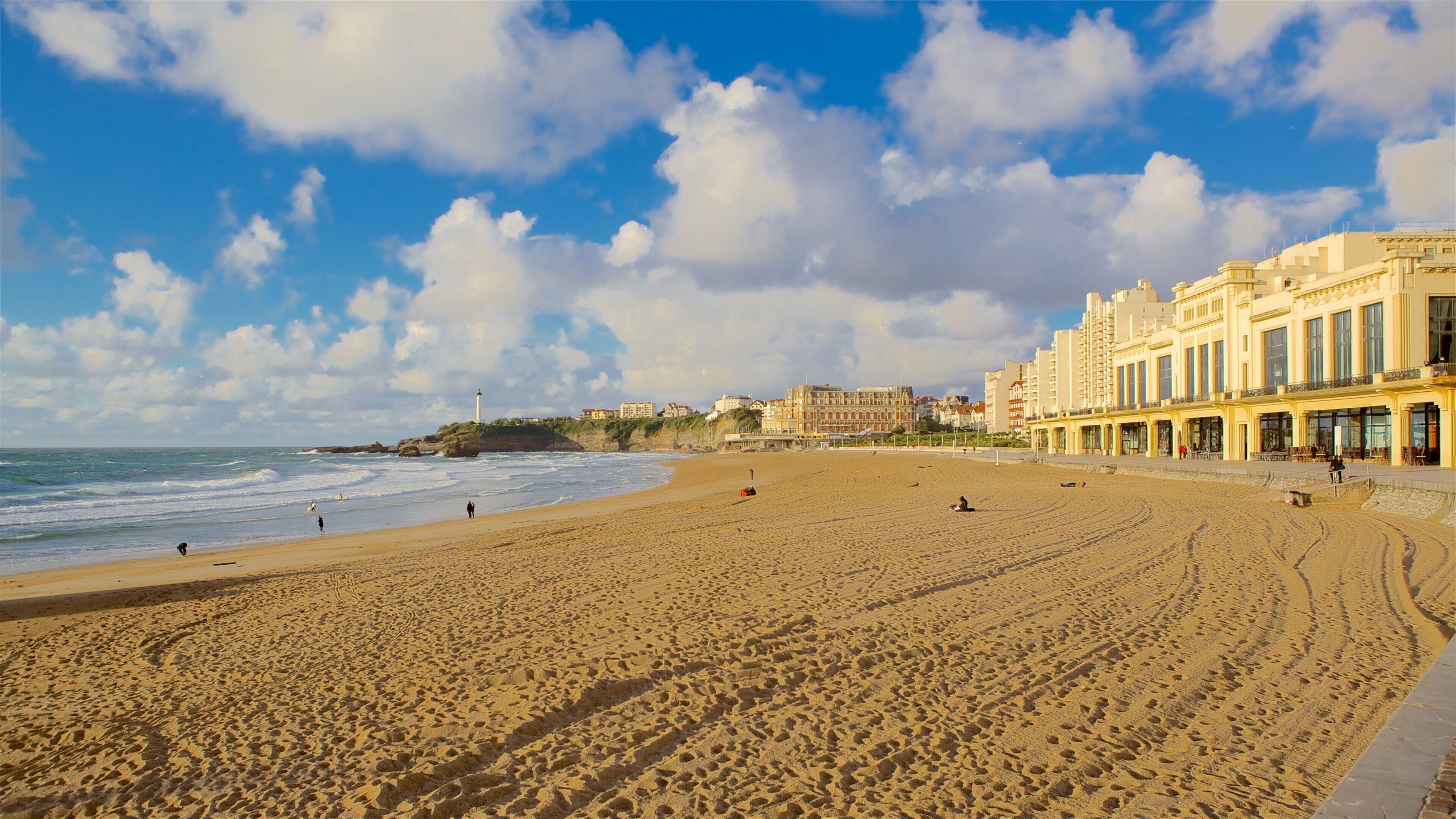 A sandy beach with footprints, a lighthouse, and buildings in the background.