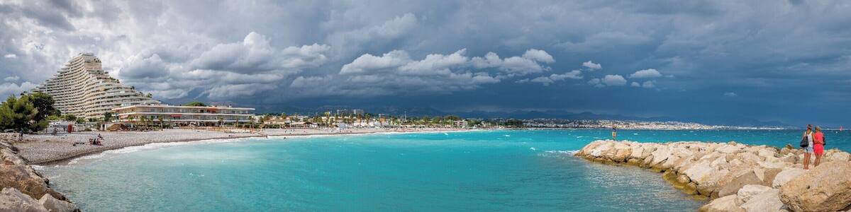 A coastal scene with a large hotel, clear turquoise waters, rocky shoreline, and a cloudy sky.