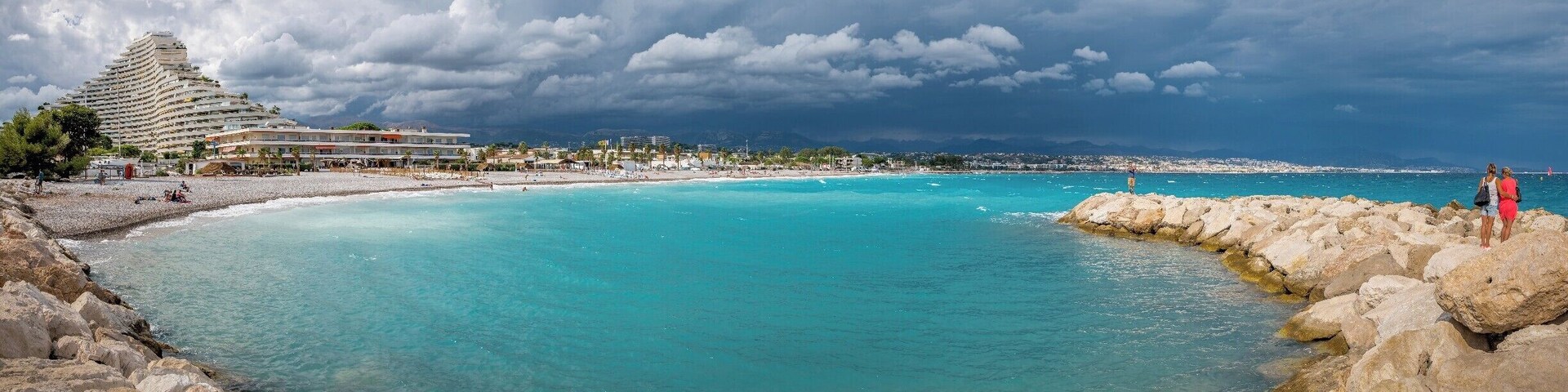 A coastal scene with a large hotel, clear turquoise waters, rocky shoreline, and a cloudy sky.
