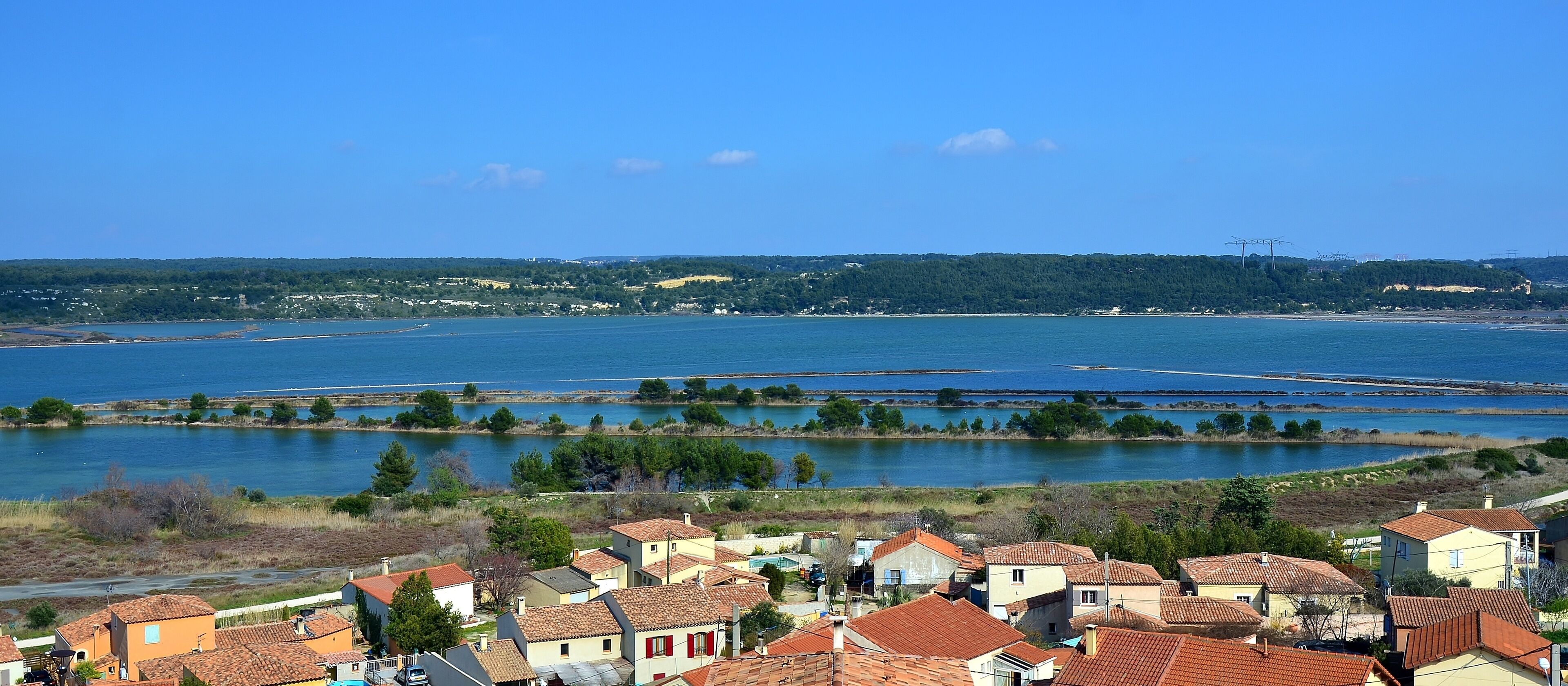 A coastal village with terracotta-roofed houses, a large body of water, and distant hills.