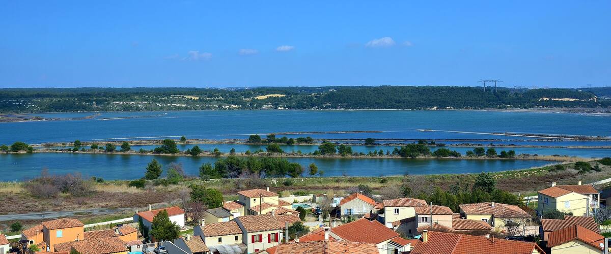 A coastal village with terracotta-roofed houses, a large body of water, and distant hills.