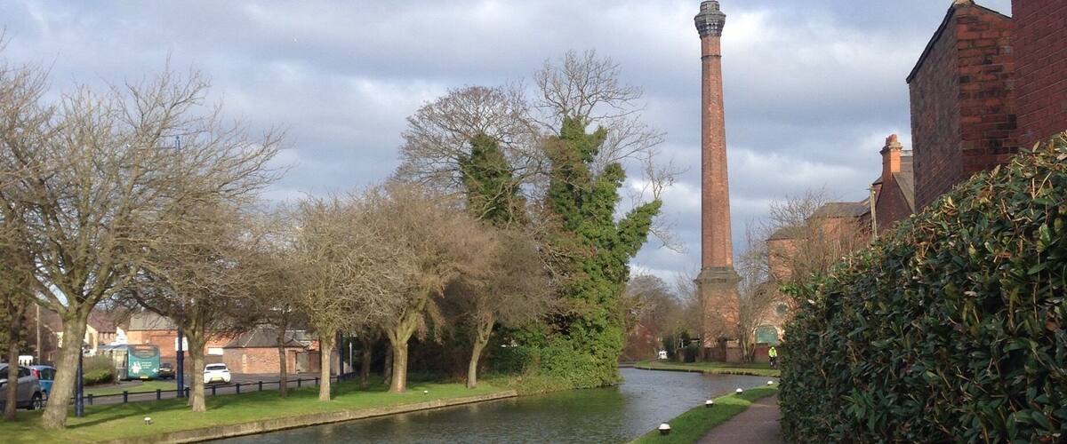 A canal with a brick tower in the distance, surrounded by trees and a hedge.